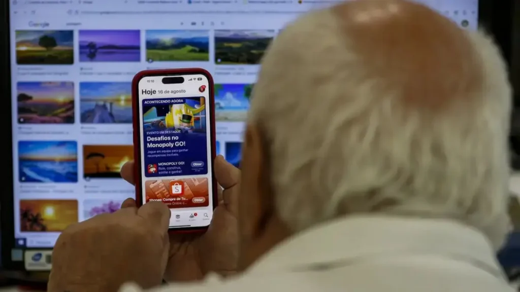 Imagem mostra idoso usando celular em frente a tela do computador