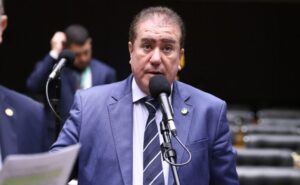 Deputado Jonas Donizette fala ao microfone. Ele é um homem claro, com cabelo escuro, veste um terno azul e camisa branca