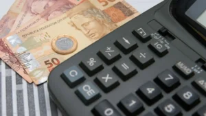 Calculadora preta sobre notas de cinquenta reais e uma moeda de um real.